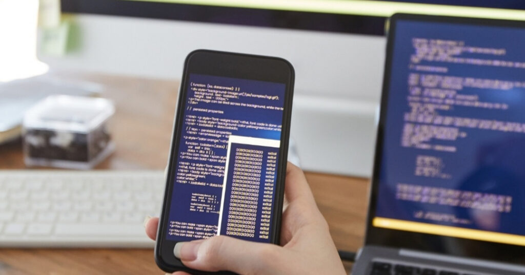 A developer holding a smartphone while working on computer code at a desk, showcasing website optimisation by Innovative Marketing SA.